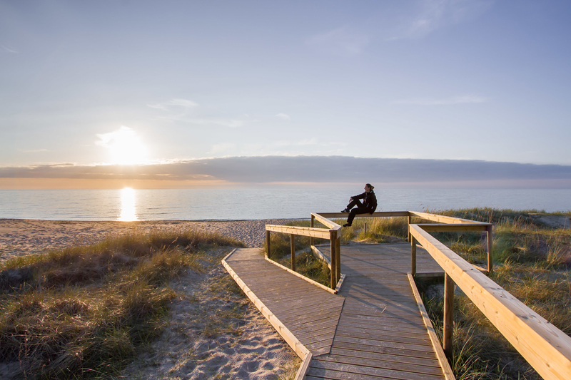 Henkilö katsoo auringonlaskua hiekkarannalla esteettömällä polulla.